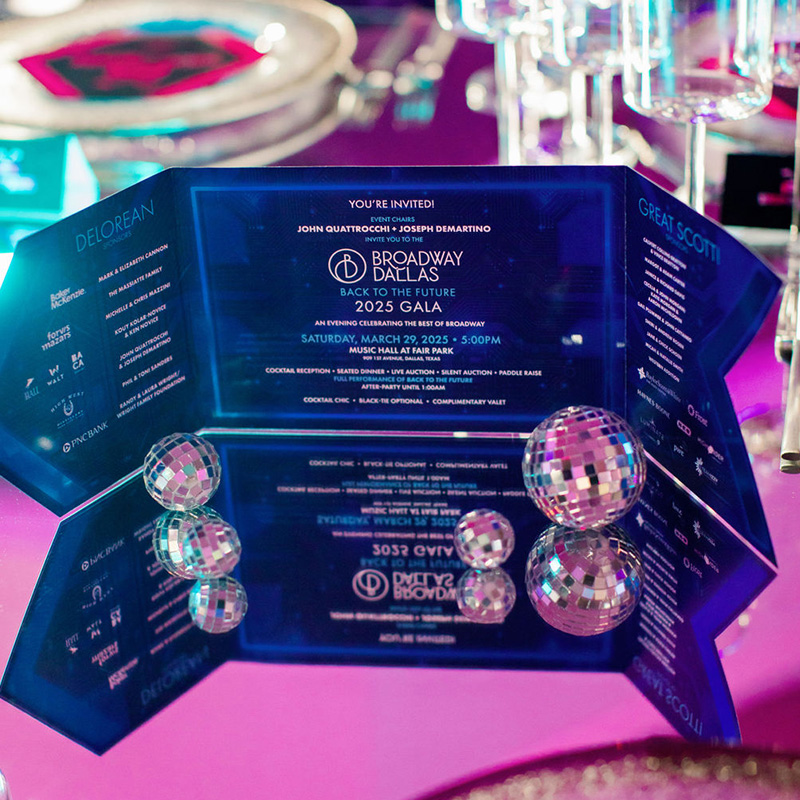A reflective, blue geometric invitation for the 2025 Broadway Gala sits on a pink surface. Small disco balls and glassware surround it, and the invitation text and design are mirrored in the surface below.
